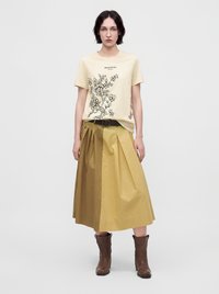 Femme portant un t-shirt crème imprimé floral, une jupe midi plissée beige bicolore avec ceinture noire et des bottines marron, debout sur un fond uni.