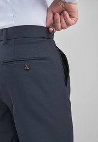 Granatowe spodnie o dopasowanym kroju, charakteryzujące się gładką fakturą, płaskim przodem oraz tylną kieszenią z zapięciem na guzik. Pętle na pasek są widoczne w talii.