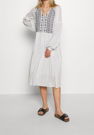 Mujer con un vestido blanco hasta la rodilla con bordado negro en el pecho y sandalias negras tipo deslizadas con hebillas.