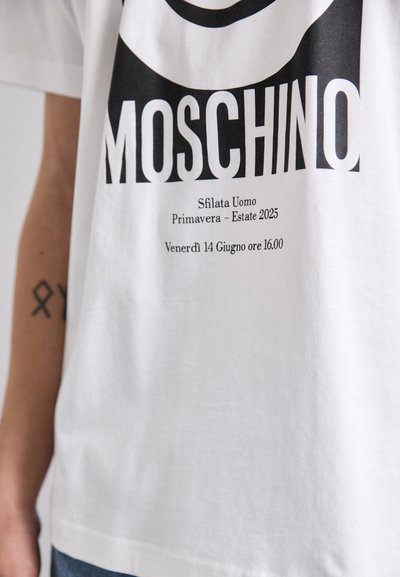 Hvit bomull t-skjorte med svart tekst. Fronten viser "Moschino" og arrangementdetaljer i fet skrift. Klassisk rund hals-design.