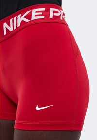 Κόκκινα σορτς Nike Pro με λευκό λογότυπο και φαρδύ ζωνάρι, φορεμένα στη λεκάνη.