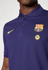 Navy polo shirt with a collar, featuring a yellow Nike swoosh, FCB badge, and Spotify logo. Made of textured fabric with two buttons.