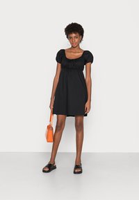 Vestido curto preto com mangas bufantes e decote quadrado. A modelo segura uma bolsa laranja. Usa sandálias pretas com sola plana.