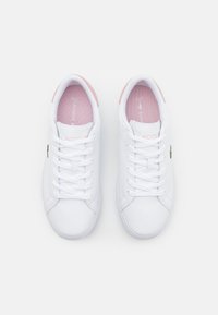 White sneakers with a smooth leather finish. Features pink accents on the heel and a lace-up design. Lacoste logo on the side.