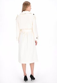 Crèmekleurige trenchcoat met een tailleband, een getailleerde pasvorm, knoopaccenten op de schouders en een midilange zoom. Gecombineerd met zwarte hoge hakken.