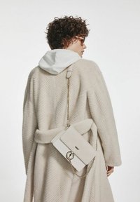 Kremowy, teksturowany płaszcz z paskiem; pod spodem biały hoodie. Na ramieniu wisi mała, biała torebka z wytłoczonym wzorem i złotym łańcuszkiem.