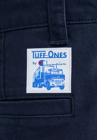 Modrá džínsovina s umístěnou nálepkou, na které je nápis "Tuff-Ones by Champion", ilustrace trucku a bílé prošívání.