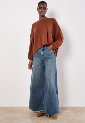 Pull oversized tricoté couleur rouille avec des manches larges associé à un jean en denim bleu taille haute et jambes larges. Texture lisse, coupe décontractée.