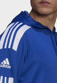 Sudadera deportiva azul con una textura suave, que cuenta con rayas blancas en los hombros y un logo bordado en el pecho.