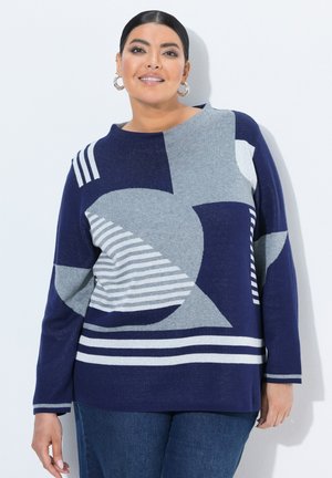 Kvinde med mørkt hår iført sølvfarvede hoops-øreringe og en marineblå og grå sweater med geometrisk mønster samt blå jeans står op ad en hvid væg.