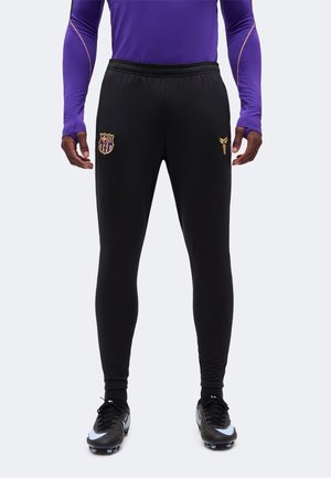 Homme portant un pantalon d'entraînement noir de football Barcelone avec des logos dorés, un haut à manches longues violet et des chaussures de football noires, debout face à l'avant.