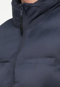 Granatowa kurtka puchowa z wysokim kołnierzem, przednim suwakiem i matowym wykończeniem. Teksturowana powierzchnia z subtelnymi przeszyciami wzdłuż szwów.