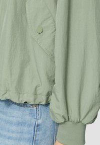 Hellgrüne, leichte Jacke mit strukturierter Oberfläche, elastischen Bündchen und einer großen aufgesetzten Tasche, die mit einem grünen Druckknopf gesichert ist.