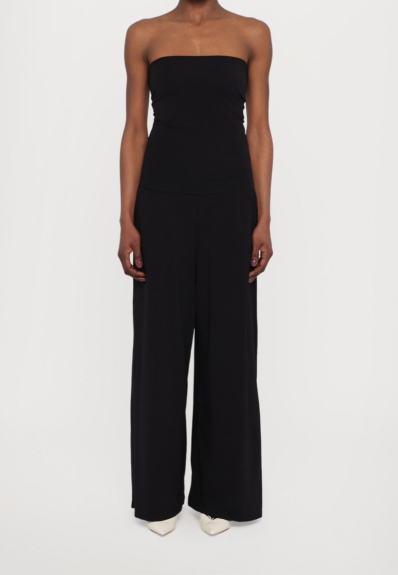 Personne portant une combinaison noire sans bretelles avec un pantalon à jambes larges et des chaussures blanches pointues, debout sur un fond blanc uni.