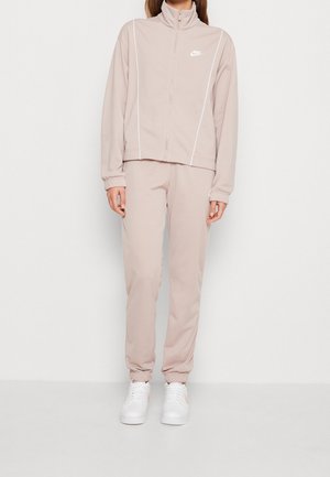 Woman wearing beige Nike tracksuit with white piping, white sneakers, standing against a plain white background.