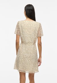 Femme aux longs cheveux foncés portant une robe beige à manches courtes avec des pois marron, vue de dos sur un fond blanc uni.
