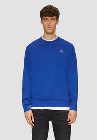 Felpa blu realizzata in misto cotone, presenta un collo rotondo, polsini a costine e un piccolo logo sul petto. Indossata sopra una maglietta bianca.