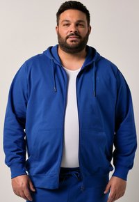 Felpa blu con zip realizzata in tessuto morbido, dotata di cappuccio con cordino, due tasche frontali e polsini a coste. Indossata sopra una maglietta bianca.