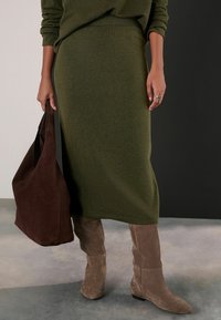 Fustă tricotată lungă, de culoare verde măslinie, cu un brâu ribbed, asortată cu cizme înalte din suede maro și o geantă tote din suede maro închis.