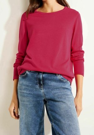 Femme portant un pull rose vif à manches longues rentré dans un jean bleu de lavage moyen, debout avec les mains détendues le long du corps.