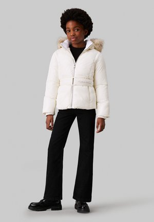 Giacca puffer bianca con cappuccio foderato in pelliccia, vita con cintura e zip argentata. Abbinata a pantaloni svasati neri e scarpe nere.