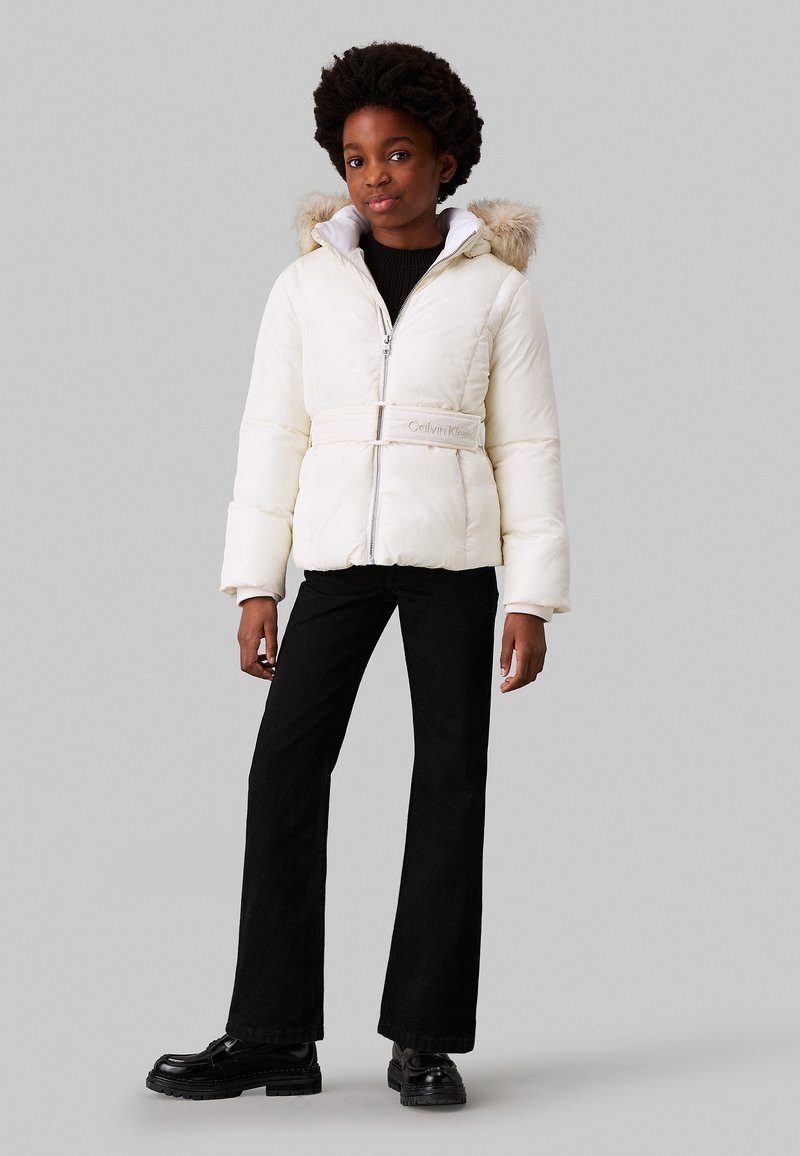 Giacca puffer bianca con cappuccio foderato in pelliccia, vita con cintura e zip argentata. Abbinata a pantaloni svasati neri e scarpe nere.