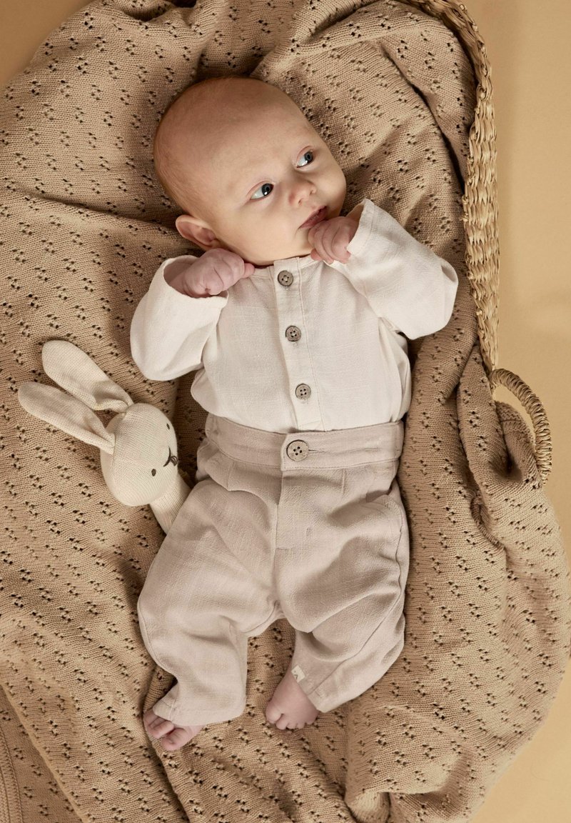 Naujagimis kūdikis dėvi smėlio spalvos aprangą, guli ant megztos antklodės pintame krepšyje šalia audinio kiškio žaislo, žiūrėdamas į šoną.