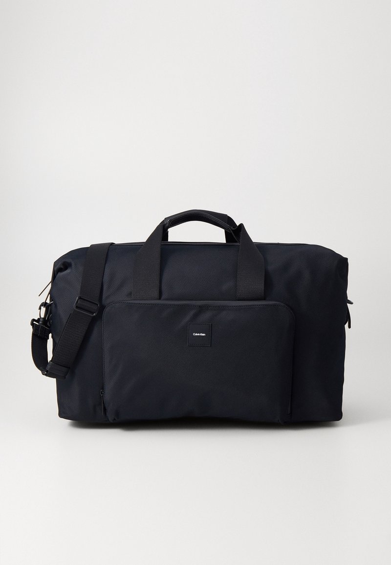Sac polochon noir en tissu durable, doté de deux poignées et d'une bandoulière amovible, avec une poche avant et un écusson marqué.