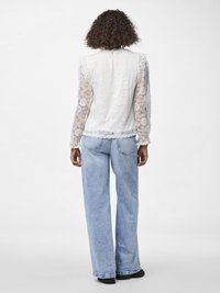 Hvit blondebluse med lange, gjennomsiktige ermer og bølgekant, kombinert med lyseblå vide jeans og svarte sko.