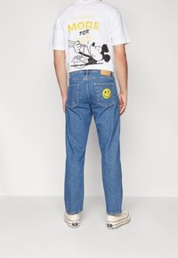 Person som bär en vit t-shirt med en Mickey Mouse-tryck och blå jeans med en gul smiley-ansikte-patch på bakfickan.