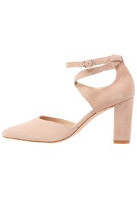 Beige mockakskinnhögklackad sko med spetsig tå, ankelrem och klack med blockform. Har en utskärningdesign på sidorna.