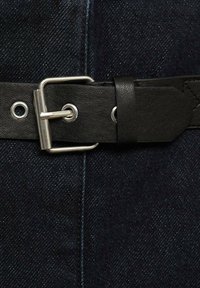Svart läderbälte med en metallspänne och öglor, placerad mot en mörk denim bakgrund, som visar sömdetaljer och textur.