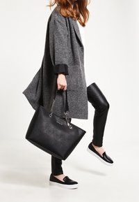 Borsa tote nera con dettagli a catena, materiale strutturato e forma definita. Indossata con un cappotto grigio oversize e pantaloni in pelle nera.