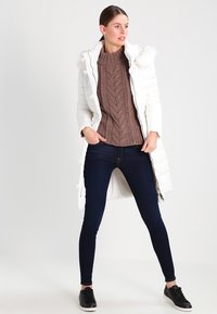 Casaco puffer branco longo com capuz, usado sobre um suéter castanho com padrão de malha e jeans skinny escuros. Ténis pretos completam o visual.