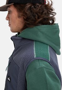 Ung man med lockigt hår, klädd i en grön huvtröja och en blå fleeceväst med dragkedja och en logotyp på bröstet, vänd mot vänster.