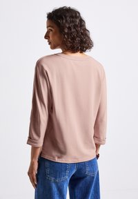 Frau mit lockigem Haar, die ein hellrosefarbenes Langarmshirt und eine blaue Jeans trägt, leicht nach links gedreht vor einem weißen Hintergrund.