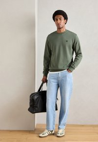 Grøn sweatshirt, lyseblå jeans og beige-stribede sneakers. Bærer en sort lædertaske. Enkel design, afslappet pasform, minimal branding.