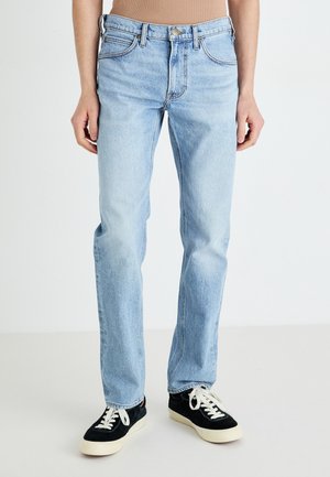 Jean en denim bleu clair avec une coupe droite, un style à cinq poches et une finition délavée, associés à des baskets noires avec des lacets blancs.