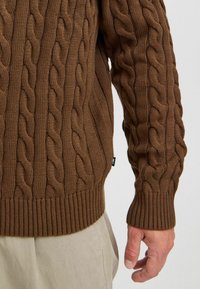 Brown cable-knit sweater featuring a ribbed hem and cuffs. The fabric has a textured appearance with intertwined patterns.