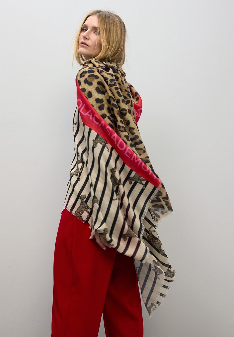 Bufanda de estampado animal y rayas con un borde rojo que presenta texto. El tejido parece ligero con bordes desflecados. Modelo llevándola sobre los hombros.