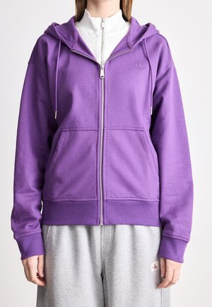 Osoba ubrana w fioletową bluzę z kapturem na zamek z przednimi kieszeniami, na białej bluzce, do tego szare spodnie dresowe z małą naszywką z logo.