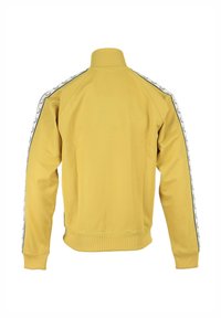 Chaqueta amarilla con cremallera y cuello alto, puños y dobladillo acanalados; presenta franjas laterales en contraste con un patrón de logo repetido.