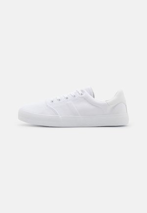 White canvas sneakers with a rubber sole, featuring six eyelets, a textured toe cap, and minimalistic design.