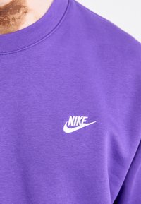 Lila pamut pulóver, amelyen egy kis fehér Nike logó található a bal mellkasi részen. Az anyag puhanak tűnik, sima textúrával.