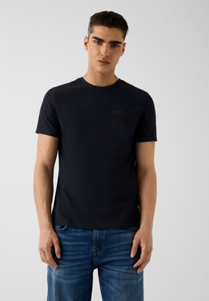 Homme aux cheveux courts et foncés portant un t-shirt noir uni à manches courtes et un jean bleu, debout devant un fond gris clair uni.