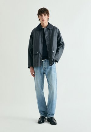 Joven con chaqueta oscura abotonada, vaqueros azul claro y zapatos negros, de pie contra un fondo claro y sencillo.