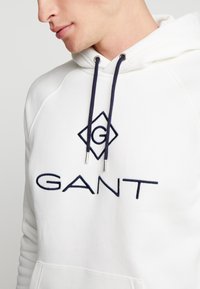 Sudadera blanca con bolsillo frontal, con un logo bordado en forma de diamante en azul marino y "GANT" en texto azul marino. Incluye cordones de ajuste en azul marino.