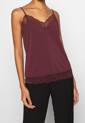 Vrouw draagt een mouwloze bordeauxrode top met kanten rand, gecombineerd met een zwarte broek, tegen een effen lichte achtergrond.