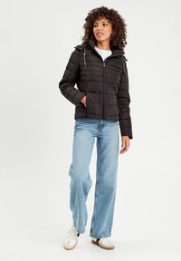 Veste matelassée noire avec un design à carreaux horizontal, fermeture éclair à l'avant, capuche ajustable et deux poches latérales. Modèle portant un jean large bleu clair et des baskets.