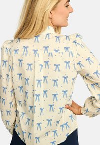 Svetlo kremna bluza z vzorcem ponavljajočih se modrih pentelj in srčkov, s poudarjenimi rokavi in zapiranjem z gumbi na hrbtu. Gladek bombažni material.